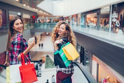 Zwei junge Frauen stehen mit Einkaufstüten vor einer Rolltreppe in einem Einkaufszentrum | © Caritas München und Oberbayern
